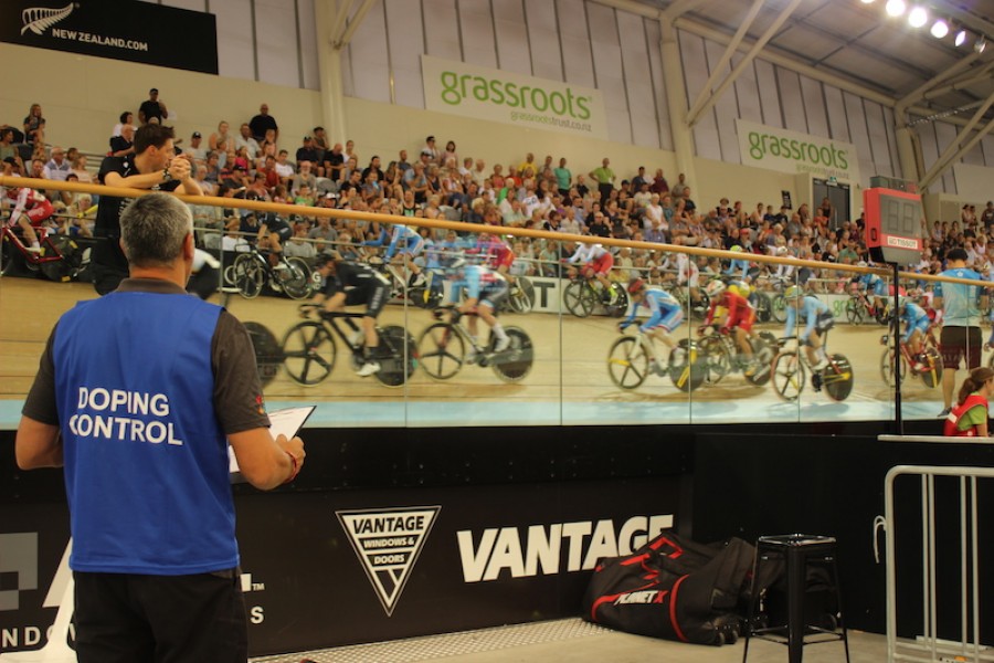 A-blur-of-cyclists-ride-past-in-an-indoor-arena-in-the-center-a-Doping-Control-Officer-looks-on..JPG