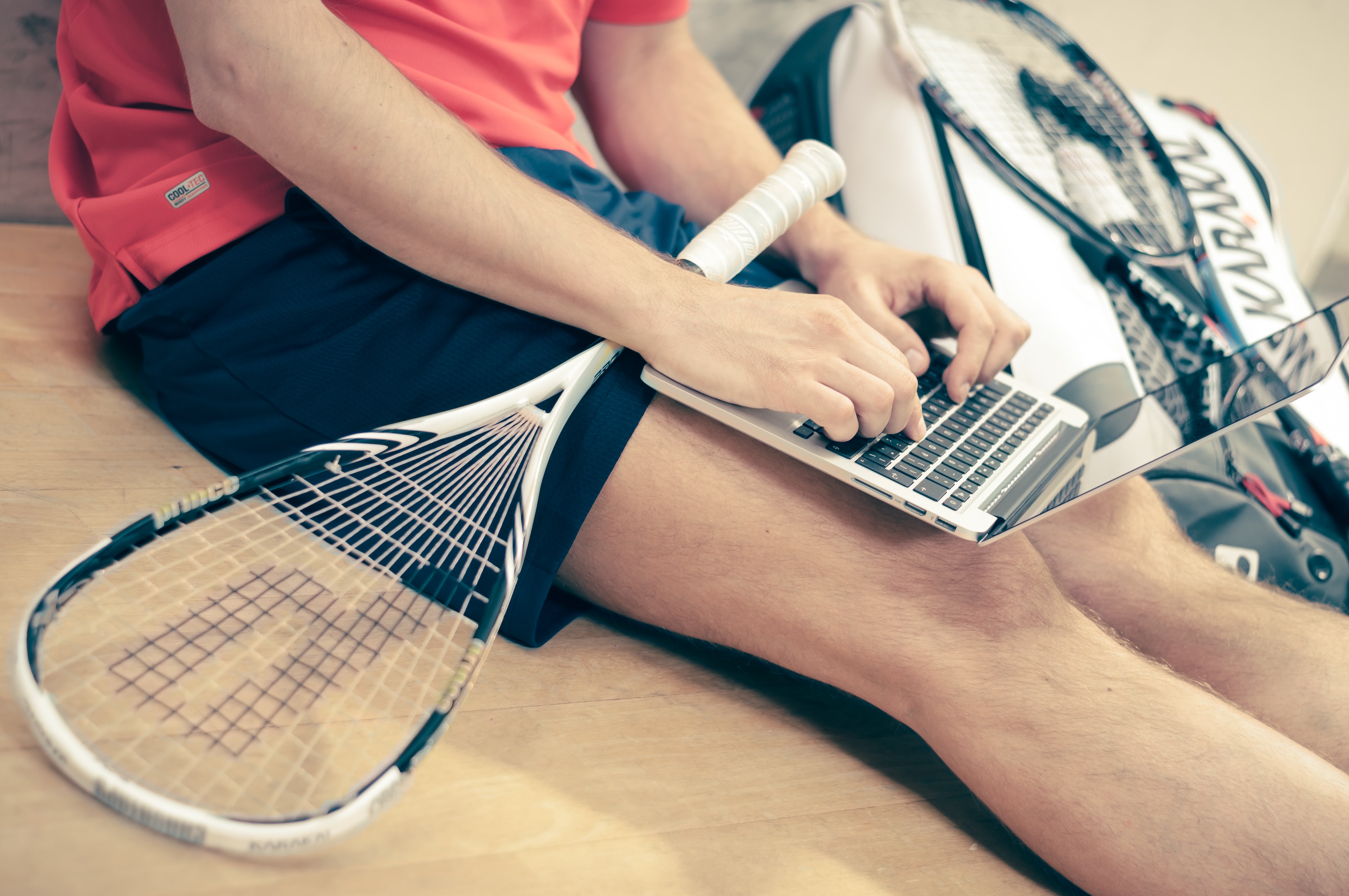Man holding squash racket sits on gym floor typing on a laptop.